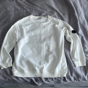 Vit sweatshirt från C.P. Company - Säljer en stilren vit sweatshirt från C.P. Company. Tröjan har en rund halsringning och långa ärmar med en snygg detalj i form av en svart logotyp på ärmen. Perfekt för en avslappnad look.