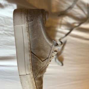 Gråa sneakers från Axel Arigato - Snygga beige sneakers från Axel Arigato i mocka med snörning. Skorna har en stilren design med en vit sula och är perfekta för en avslappnad look.