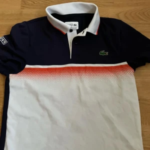 Svart och vit pikétröja från Lacoste - Snygg pikétröja från Lacoste i svart och vitt med en cool orange detalj. Tröjan har en klassisk krage och knappar framtill. Perfekt för en stilren look.