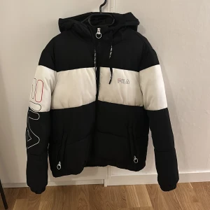 Svart och vit pufferjacka från Fila - Snygg svart och vit pufferjacka från Fila med dragkedja och huva. Jackan har ett stort Fila-tryck på ärmen och är perfekt för kyligare dagar. Den har en sportig look och är både stilren och bekväm.