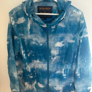 Woolrich windbreaker  - Säljer en cool blå mönstrad jacka från Woolrich. Jackan har en unik design med ett abstrakt mönster i blått och vitt. Den är långärmad och har en dragkedja framtill. Perfekt för att sticka ut i mängden!