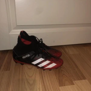 Adidas Predator fotbollsskor - Snygga Adidas Predator fotbollsskor i svart och rött med vita detaljer. Skorna har snörning och en hög ankelkrage för extra stöd. Perfekta för fotbollsplanen med sina greppvänliga dobbar.