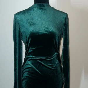 Grön sammetsklänning - Elegant grön sammetsklänning med långärmad design och en åtsittande passform. Klänningen har en subtil glans och rynkade detaljer som ger en lyxig känsla. Perfekt för speciella tillfällen.