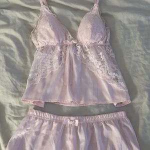 Rosa spetslinne och shorts - Säljer ett sött rosa set med spetslinne och matchande shorts. Linnet har justerbara axelband och spetsdetaljer med en liten rosett framtill. Shortsen har en elastisk midja och spetskant. 