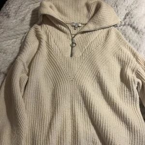 Beige stickad tröja från b.young - Säljer en mysig beige stickad tröja från b.young. Tröjan har en dragkedja framtill och en hög krage som ger extra värme. Perfekt för kyliga dagar!