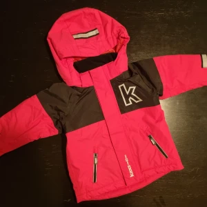 Rosa och svart vinterjacka från Kaxs - Mycket fin ny snygg rosa och svart vinterjacka från Kaxs i storlek 92. Jackan har en huva, fickor med dragkedja och reflexdetaljer för extra synlighet. Den är tillverkad i nylon med polyesterfoder.. Perfekt för kyliga dagar!