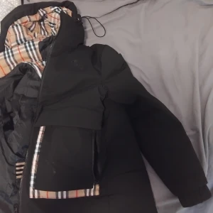 Svart dunjacka från Burberry - Säljer en stilren svart dunjacka från Burberry med klassiskt rutigt foder. Jackan har en dragkedja framtill och en huva för extra skydd mot väder. Perfekt för kyliga dagar med sin varma och bekväma design.