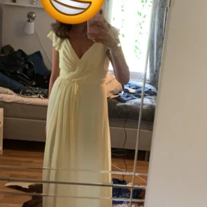Ljusgul långklänning med volangdetaljer - Säljer min fina balklänning som har en fin ljusgul färg med v-ringning och volangdetaljer vid axlarna. Klänningen har en knytning i midjan som ger en smickrande passform. Den är endast använd en gång och det finns inga tecken på tidigare användning!🩷