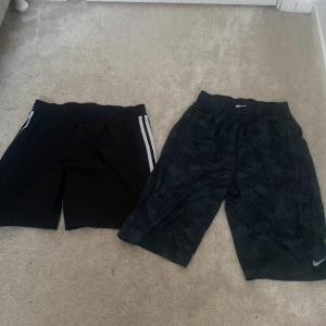 Svarta träningsshorts från Nike och Adidas  - Två par svarta träningsshorts, ett från Nike med Dri-FIT-teknologi och ett från Adidas. Nike-shortsen har en diskret logga och elastisk midja. Adidas-shortsen har en klassisk design med vita ränder på sidorna. Perfekta för sport och träning. Nike är XL och adidas är M