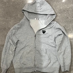 Zip hoodie COMMES DES GARCONS - Säljer en grå hoodie från Play med dragkedja och en stor svart hjärtdetalj med ögon på ryggen. Framtill finns en mindre hjärtdetalj på bröstet. Perfekt för en avslappnad stil.
