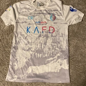 Vit fotbollströja från Al Nassr - Snygg vit fotbollströja från Al Nassr med CR7-tryck och nummer 7 på ryggen. Tröjan har korta ärmar och flera sponsorloggor i olika färger på framsidan. Perfekt för fotbollsfans!