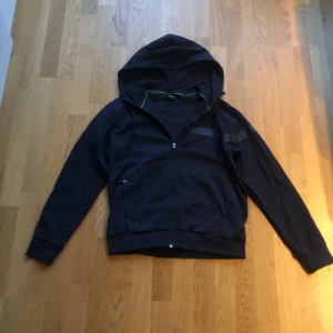 Mörkblå huvtröja från Boss - Snygg börkblå huvtröja från Boss med dragkedja och gröna detaljer. Perfekt för en casual look med sin bekväma passform och stilrena design. Huvan ger extra skydd och stil. Storlek, S. Nästan oanvänd. Köpt för 2200 ungefär på pondus i Partille/allum.