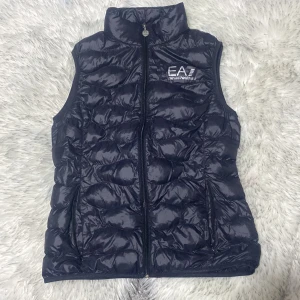 EA7 väst  - Snygg svart dunväst från Emporio Armani med dragkedja och hög krage. Perfekt för kyligare dagar och ger en stilren look. Västen har en quiltad design och en vitt EA7-logga på bröstet.