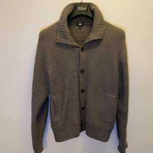 Brun cardigan - Säljer en stilren cardigan från Uniqlo med ribbad design och knappar framtill. Koftan har en hög krage och långa ärmar, perfekt för kyligare dagar. Den har också praktiska fickor på sidorna.