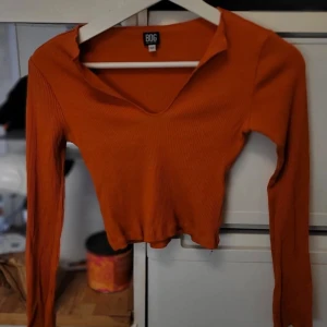 Orange ribbad tröja från BDG - Säljer en snygg orange ribbad tröja från BDG. Tröjan har en v-ringad design och långa ärmar, perfekt för en trendig look. Den är croppad och passar bra till både jeans och kjol. Perfekt för höstgarderoben!