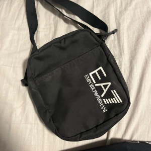 Svart axelväska från Emporio Armani - Snygg svart axelväska från Emporio Armani med justerbar rem. Väskan har en praktisk dragkedja och logotypen tryckt på framsidan. Perfekt för att bära med sig det nödvändigaste med stil.