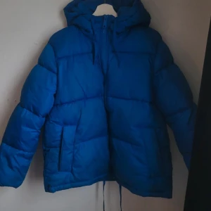 Blå pufferjacka med huva - Säljer en blå pufferjacka med huva. Jackan har långa ärmar och dragsko i nederkant för justerbar passform. Perfekt för kyliga dagar.