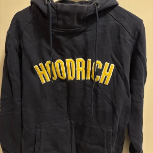 Mörkblå hoodie från Hoodrich - Säljer en snygg mörkblå hoodie från Hoodrich med gul text på framsidan. Den har en stor ficka framtill och justerbar huva med snören. Perfekt för en avslappnad stil.
