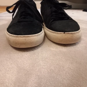 Axel Arigato skor - Snygga svarta sneakers från Axel Arigato med vit sula och diskret logga på sidan. Skorna har snörning och är tillverkade i mocka och skinn, vilket ger en stilren look. Med 1 liten defekt.