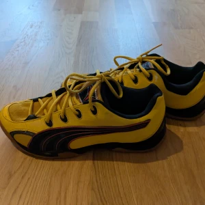 Gula sneakers från Puma - Snygga gula sneakers från Puma med svarta och röda detaljer. Skorna har en klassisk snörning och en sportig design. Perfekta för en casual look med en touch av färg.