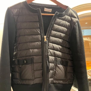 Svart cardigan från Moncler - Snygg svart jacka från Moncler med quiltade detaljer och dragkedja framtill. Jackan har långa ärmar och praktiska fickor med knappar. Storlek XS