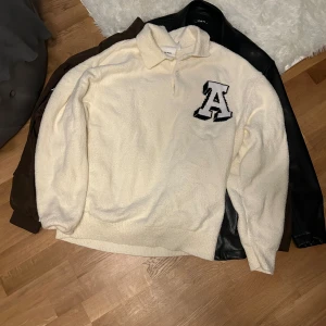 Beige pikétröja med bokstavsdetalj - Säljer en beige pikétröja med en stor svart bokstav 'A' på bröstet. Tröjan har en klassisk krage och knappar framtill. Perfekt för en avslappnad stil.