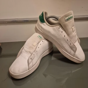 Vita Adidas Stan Smith sneakers - Klassiska vita Adidas Stan Smith sneakers med gröna detaljer på hälen. Skorna har en stilren design med perforerade ränder på sidorna och en bekväm sula. Perfekta för en avslappnad stil.
