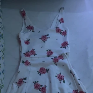 Vit klänning med rosor - Charmig vit klänning med rosa blommönster. Klänningen har smala axelband och en utsvängd kjol som ger en feminin siluett. Perfekt för somriga dagar.