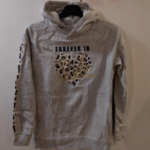 Grå hoodie med leoparddetaljer - Säljer en grå hoodie med texten 'FOREVER IN' och ett leopardmönstrat hjärta på framsidan. Ärmarna har leopardmönstrade detaljer längs sidorna. Perfekt för en avslappnad stil.