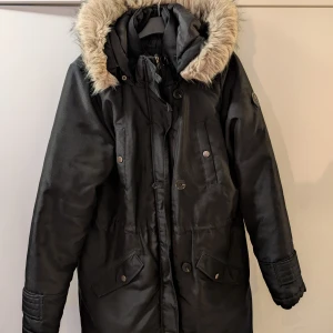 Svart parkajacka med pälsfodrad huva - Snygg svart parkajacka med pälsfodrad huva och knappar. Jackan har flera praktiska fickor och en stilren design. Perfekt för kyliga dagar.
