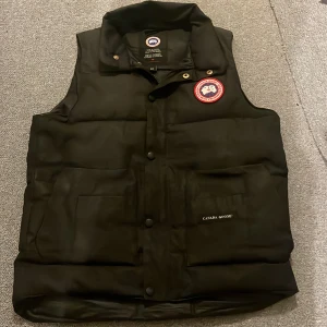 Svart dunväst från Canada Goose - Säljer en svart dunväst från Canada Goose med knappar och broderad logotyp på bröstet. Västen har en klassisk design med praktiska fickor och hög krage för extra värme. Perfekt för kyliga dagar.