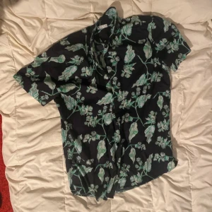 Svart blommig skjorta - Snygg svart skjorta med grönt blommönster. Perfekt för en avslappnad stil med korta ärmar och knappar framtill. Passar bra till både jeans och shorts. Använda endast en gång