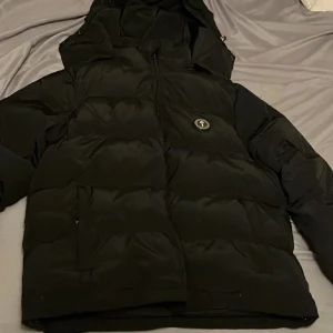 Svart pufferjacka från Trapstar  - Snygg svart pufferjacka från Trapstar med huva och dragkedja. Jackan har ett broderat Trapstar -märke på bröstet och text på ryggen. Perfekt för kalla dagar. Luvan går att ta av om man så vill.             Passar bra till storlek L 