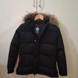 Svart dunjacka från Canada Goose - Säljer en svart dunjacka från Canada Goose med pälskantad huva. Jackan har en klassisk design med långärmade ärmar och en broderad logotyp på ärmen. Perfekt för kalla vinterdagar.