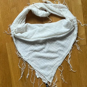 Vit halsduk med fransar - Säljer en stilren vit halsduk med fransar. Den har ett diskret rutmönster och är perfekt för att ge en touch av elegans till din outfit. Passar bra för svalare dagar.