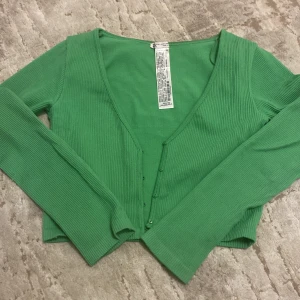 Grön ribbad kofta från Zara - Säljer en snygg grön ribbad kofta från Zara med knappar framtill. Perfekt för lager-på-lager och att hålla stilen året runt. Koftan har långa ärmar och en v-ringad design.
