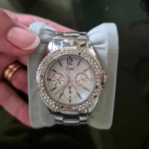 Silverfärgad klocka från Guess - Elegant silverfärgad klocka från Guess med glittrande stenar runt urtavlan. Klockan har en vit urtavla med tre mindre visare för extra funktioner. Perfekt accessoar för att lyfta din stil. Tappat några stenar därav det låga priset.