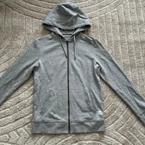 Grå hoodie HELT NY - HELY NYTT, Säljer en stilren grå hoodie  med dragkedja och justerbar huva. Perfekt för en avslappnad look. Den har praktiska fickor framtill och är tillverkad i ett mjukt material. 