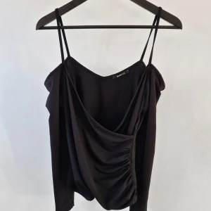 Svart topp från Gina Tricot - Snygg svart topp från Gina Tricot med smala axelband och draperad design. Perfekt för en stilren look. Toppen har en elegant passform och är tillverkad i ett mjukt material.