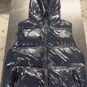 Svart glansig dunjacka från Moncler (barn) - Säljer en svart maya dunjacka från Moncler med huva och dragkedja. Jackan har en quiltad design och är perfekt för kyligare väder. Den är dock skadad på en av fickorna och på insidan men man tänker ej på det