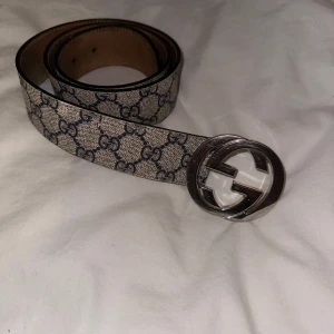 Mönstrat bälte från Gucci - Snyggt bälte från Gucci med klassiskt mönster och en iögonfallande silverfärgad spänne. Bältet har en beige och blå färgkombination som ger en tidlös look. Perfekt för att lyfta vilken outfit som helst.