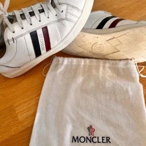 Vita sneakers från Moncler - Snygga vita sneakers från Moncler med klassiska ränder i blått och rött på sidan. Skorna har snörning och är tillverkade i skinn. Perfekta för en stilren look.