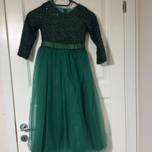Grön glittrig klänning med tyllkjol - Elegant grön klänning med glittrig överdel och lång tyllkjol. Klänningen har trekvartsärmar och ett fint knytband i midjan. Perfekt för festliga tillfällen.