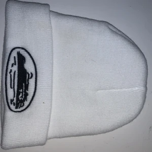 Vit stickad mössa med broderad logga - Snygg vit stickad mössa med en broderad svart logga framtill. Perfekt för att hålla värmen under kyliga dagar. Enkel och stilren design som passar till det mesta.