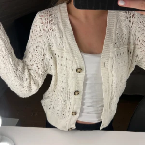 Vit stickad kofta med knappar - Säljer en söt vit stickad kofta med dekorativa knappar framtill. Koftan har ett fint hålmönster och långa ärmar, perfekt för lager-på-lager-stil. Passar bra till både jeans och kjol.