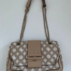 Beige axelväska från Guess - Snygg beige axelväska från Guess med kedjedetaljer och ett elegant mönster. Väskan har en praktisk dragkedja och stiliga nitar på framsidan. Perfekt för att ge din outfit en lyxig touch.
