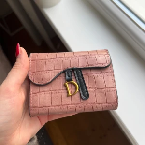 Rosa plånbok  - Snygg rosa plånbok med krokodilmönster och guldfärgad D-detalj. Plånboken har en svart kant och en praktisk knappstängning. Insidan är i en kontrasterande orange färg med plats för kort.