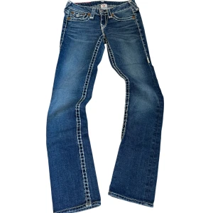 Blå lågmidjade jeans från True Religion - Snygga blå  jeans från True Religion med kontrastsömmar och klassisk femficksdesign. Jeansen har en låg midja och är perfekta för en avslappnad stil. Märkeslogga på bakfickan ger en extra touch.