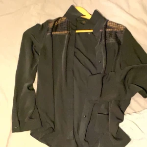 Svart långärmad skjorta - Snygg svart långärmad skjorta med knappar och diskreta detaljer på axlarna. Perfekt för en stilren look.