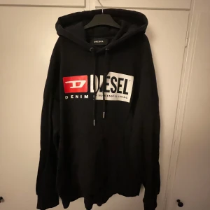 Svart hoodie, Diesel - Snygg svart hoodie med stor logga i vitt och rött på framsidan. Den har en klassisk passform med långa ärmar och justerbar huva med snören. Perfekt för en avslappnad stil.